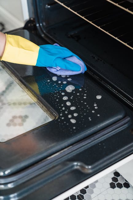 Close-up of a person wearing a yellow and blue cleaning glove, applying surface cleaning to a black ceramic hob on an oven, with visible soap suds and bubbles on the stovetop surface. The oven interior, with metal racks, is open and appears clean. The countertop surrounding the oven has a tiled surface featuring black and white geometric patterns, and the lighting highlights the shiny, well-maintained surfaces. This scene demonstrates an attentive approach to deep cleaning and sanitisation of domestic kitchen appliances, showcasing thorough maintenance by Haringey Cleaners in Haringey N15 as part of their professional cleaning service category.