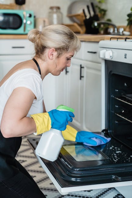 A woman with blonde hair tied in a bun wearing a white t-shirt, yellow and blue cleaning gloves, is kneeling in front of an open black oven in a modern kitchen. She is holding a spray bottle in her right hand and a blue cleaning cloth in her left, appearing to clean the oven's interior surface, which shows some dust and residue. The kitchen features a wooden countertop with various kitchen utensils, a microwave, and a potted plant. The oven's stainless steel door is partially open, revealing the oven rack inside, and the overall scene depicts surface cleaning and maintenance as part of domestic cleaning services. The setting is well-lit, emphasizing cleanliness and hygiene, with a focus on thorough oven and kitchen surface cleaning, as offered by Haringey Cleaners.
