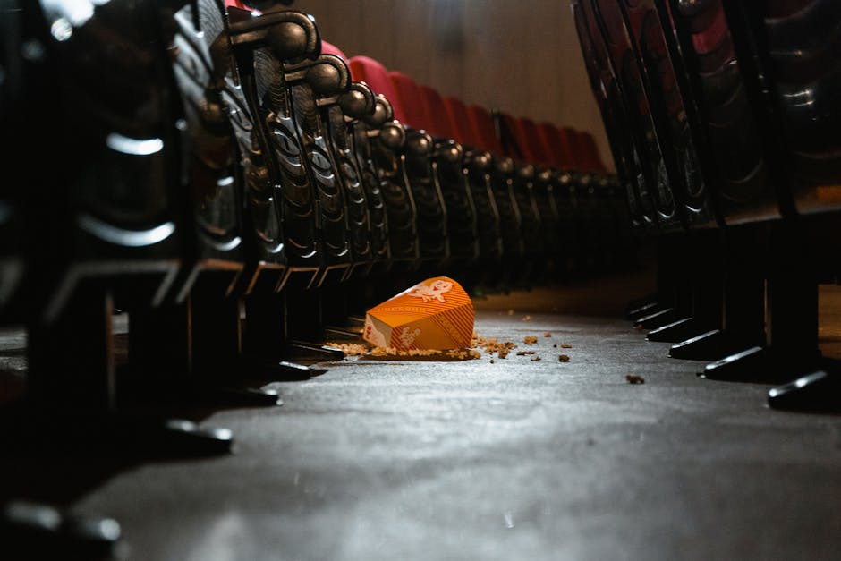 A close-up view of an auditorium or conference hall floor with black stackable chairs arranged along a central aisle. The floor surface appears to be carpeted or made of dark material, showing some scattered debris and crumb residues, including an orange and white popcorn box tipped over in the middle of the aisle. The lighting creates a slight sheen on the floor, emphasizing the cleanliness level, while the chairs display a neat, orderly arrangement. The scene suggests recent event cleanup activity, with visible evidence of debris needing removal. The image reflects the importance of surface cleaning and sanitisation for venue hygiene, and it features the tools and environment associated with professional cleaning services like Haringey Cleaners, who specialise in venue cleaning and event space maintenance.