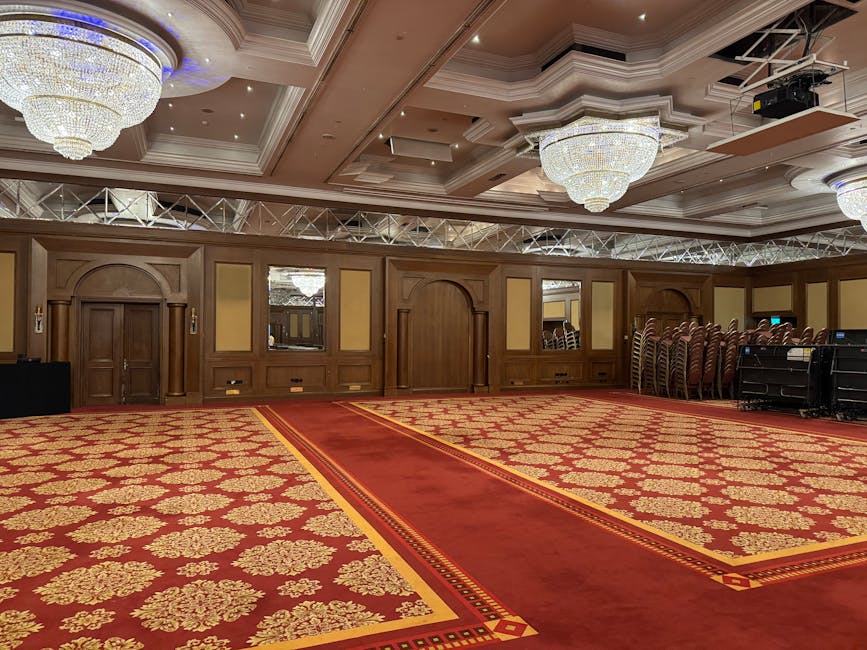 A spacious banquet hall with a high ceiling featuring ornate crown molding and three large, elegant chandeliers emitting bright light. The room has a patterned red and gold carpet covering the entire floor, which appears clean and well-maintained. On the back wall, there are wooden paneling with arched doorways and decorative mirrors, adding a sophisticated touch. To the right, there is a stack of chairs and equipment carts, indicating recent or upcoming venue cleaning. The lighting highlights the polished surfaces and the tidy, dust-free condition of the space, reflecting thorough surface cleaning and sanitisation by Haringey Cleaners as part of their event cleanup and venue cleaning services in Alexandra Palace, Haringey.
