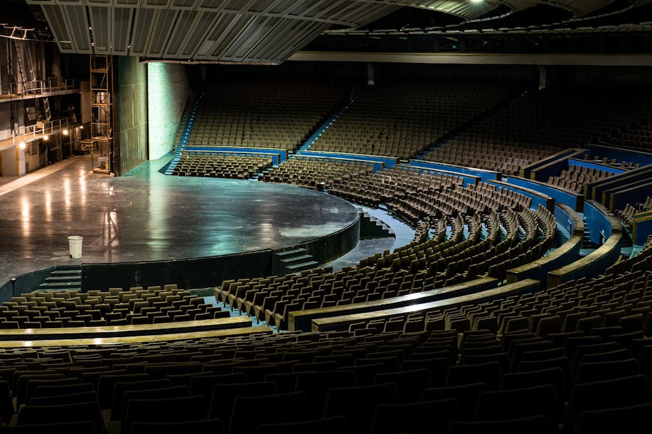 A close-up view of an auditorium or conference hall floor with black stackable chairs arranged along a central aisle. The floor surface appears to be carpeted or made of dark material, showing some scattered debris and crumb residues, including an orange and white popcorn box tipped over in the middle of the aisle. The lighting creates a slight sheen on the floor, emphasizing the cleanliness level, while the chairs display a neat, orderly arrangement. The scene suggests recent event cleanup activity, with visible evidence of debris needing removal. The image reflects the importance of surface cleaning and sanitisation for venue hygiene, and it features the tools and environment associated with professional cleaning services like Haringey Cleaners, who specialise in venue cleaning and event space maintenance.