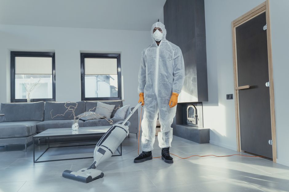 A professional cleaner dressed in a full white protective suit, face mask, and orange gloves is operating a modern vacuum cleaner on a light grey tiled floor in a contemporary living room. The room features a grey fabric sofa with cushions, a black coffee table, and two large windows allowing natural light to illuminate the space. The background includes a dark wall with a built-in fireplace, and a closed door with a wooden frame. The scene emphasizes hygienic surface cleaning and deep cleaning processes essential for maintaining a sanitized residential environment, consistent with the services offered by Haringey Cleaners to protect against hidden cleaning charges in Haringey, as detailed on the webpage.