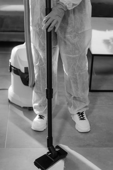 A person wearing protective gloves and a disposable suit is using a handheld vacuum cleaner with a long, extendable hose to perform surface cleaning on a tiled floor in an indoor setting. The vacuum has a flat, rectangular head that is making contact with the floor, which appears to be smooth ceramic tile, clean and dry. In the background, there is a white portable cleaning machine or extractor with a hose attached, placed near a dark piece of furniture or wall. The area is well-lit, emphasizing the cleanliness of the surface. This image illustrates professional deep cleaning or sanitisation procedures, aligning with domestic cleaning services offered by Haringey Cleaners, highlighting the importance of maintaining hygienic indoor surfaces to prevent hidden cleaning charges, as discussed in the article 'Protect against hidden cleaning charges in Haringey what to know'.