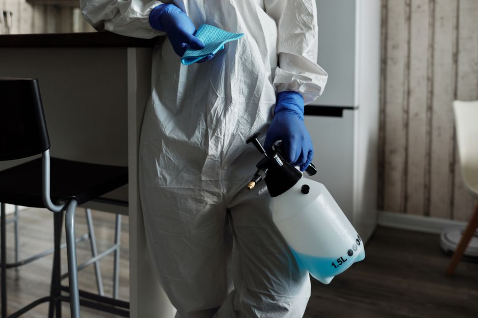A professional cleaner dressed in a full white protective suit, face mask, and orange gloves is operating a modern vacuum cleaner on a light grey tiled floor in a contemporary living room. The room features a grey fabric sofa with cushions, a black coffee table, and two large windows allowing natural light to illuminate the space. The background includes a dark wall with a built-in fireplace, and a closed door with a wooden frame. The scene emphasizes hygienic surface cleaning and deep cleaning processes essential for maintaining a sanitized residential environment, consistent with the services offered by Haringey Cleaners to protect against hidden cleaning charges in Haringey, as detailed on the webpage.