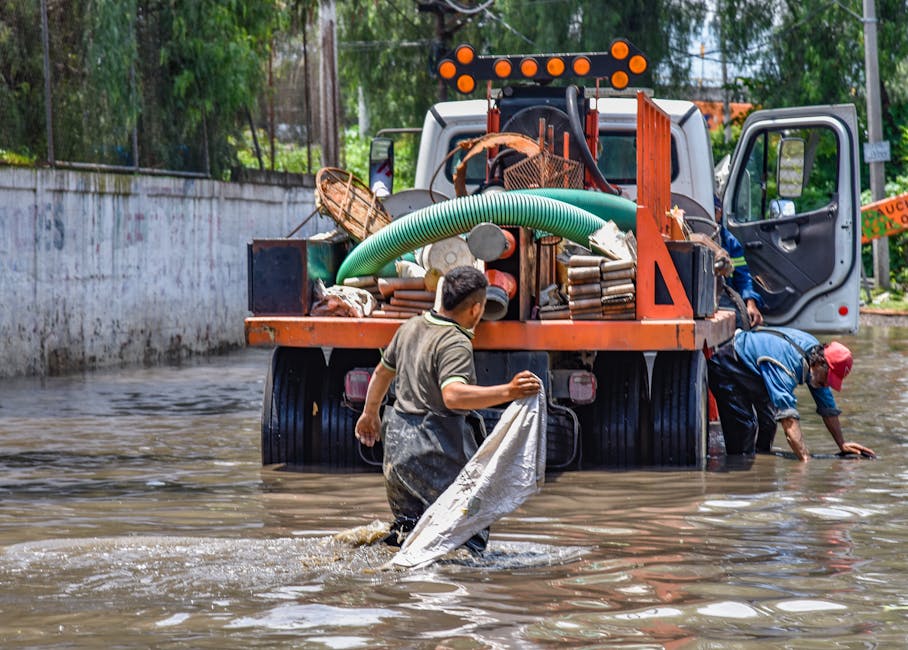 Two workers stand knee-deep in muddy floodwater next to a large orange flood response truck. One worker, wearing a gray shirt and waterproof pants, holds a white cloth or tarp, while the other, dressed in a blue jacket and red cap, bends down retrieving items from the submerged ground. The truck is equipped with hoses, tools, and supplies for flood remediation, positioned on a flooded street with a concrete wall and trees in the background. The scene illustrates active flood cleanup efforts, with dim lighting highlighting the wet surfaces and muddy water. The team from Haringey Cleaners is engaged in surface cleaning and flood damage mitigation, demonstrating emergency response procedures to restore hygiene and safety in affected areas.