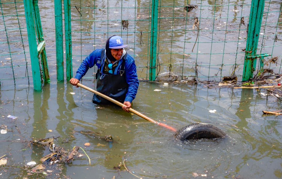 A person wearing a blue jacket, blue cap, and rubber gloves is standing in thigh-high floodwater in an outdoor area, using a long wooden stick to remove debris and clear a submerged tire near a green metal fence. The water appears muddy with scattered litter and plant material, and the scene is illuminated by natural daylight. The area looks affected by flooding, necessitating surface cleaning and debris removal. The image depicts emergency flood response efforts, consistent with the urgent flood cleaning services provided by Haringey Cleaners, as featured on the page titled 'Urgent flood cleaning in Tottenham Hale Haringey N17' at haringeycleaners.org.uk.